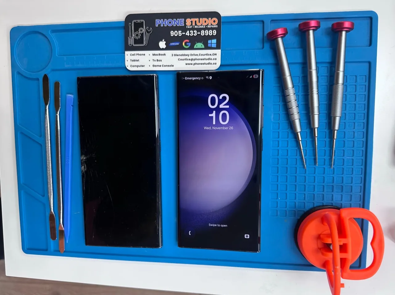 Samsung phone screen replacement: a damaged panel beside the working phone on a blue repair mat at Phone Studio